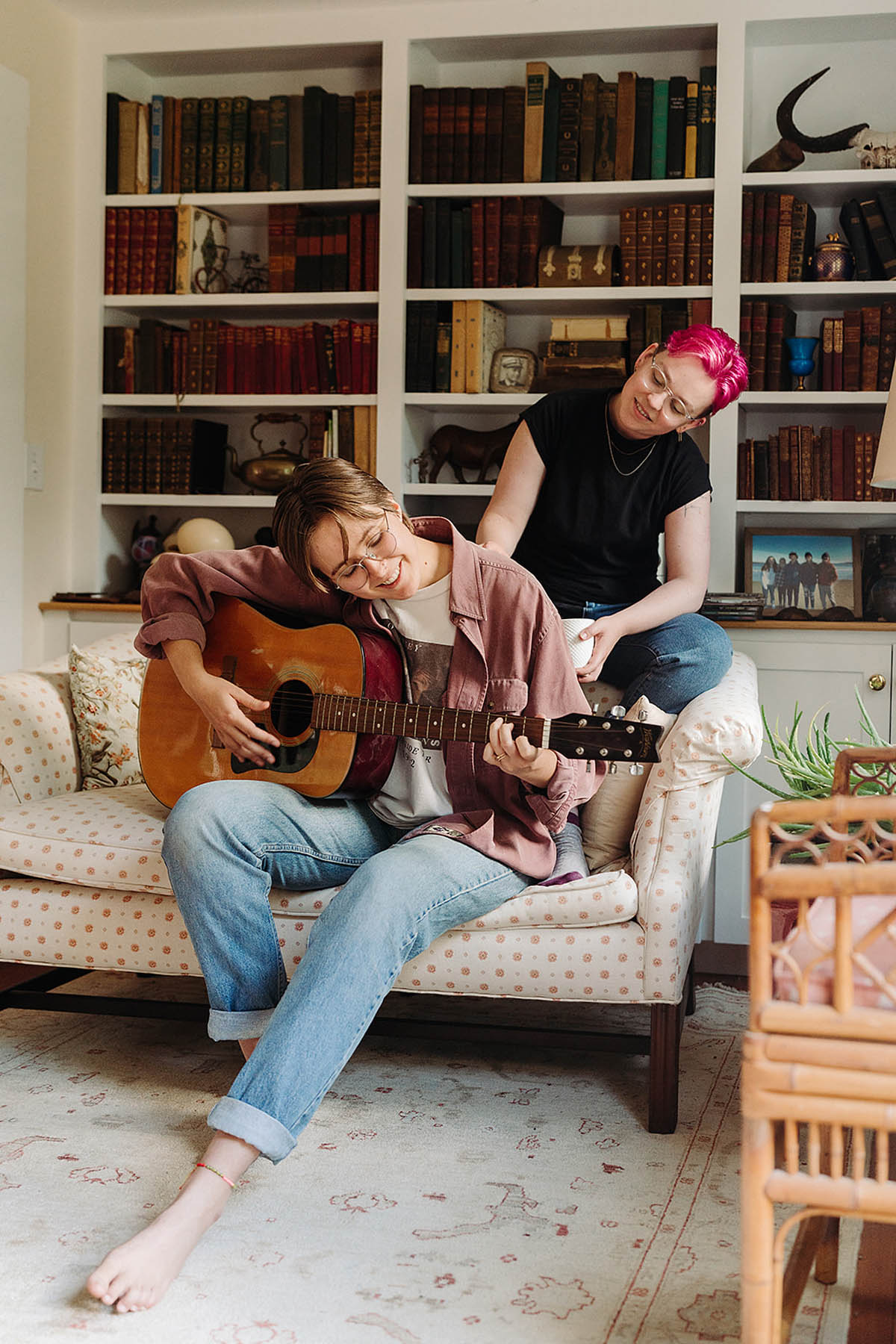 Two white people sit on a yellow sofa posing for documentary-style engagement photos. One person has pink hair and is wearing a black shirt, and the other is a brunette wearing blue jeans and playing a guitar.