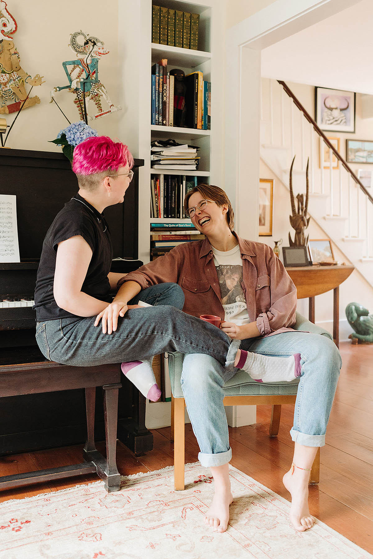 Two white people sit in their living room, one perched on a piano bench with their legs in the other's lap. The other person is sitting in a chair, and both are smiling at each other. 