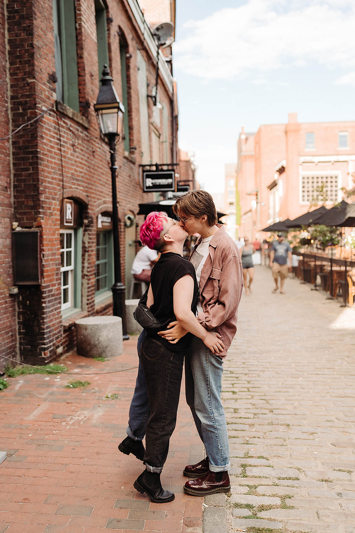 Two white people stand kissing on a red brick sidewalk in Maine. The one on the left has pink short hair, and the other has short brown hair.