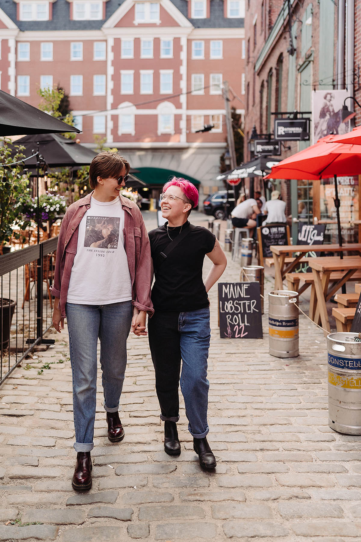 Two white people hold hands as they walk down a cobblestone street. There is a sign behind them that says "Maine Lobster Roll $22"