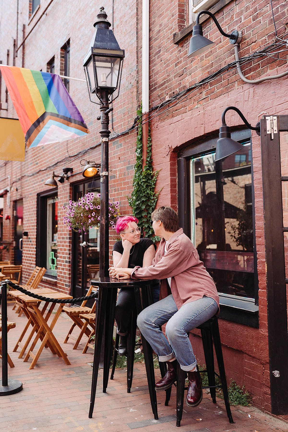 Two white people sit at a high-top table outside a restaurant. They are looking at each other smiling. Behind them there is a Pride flag. 