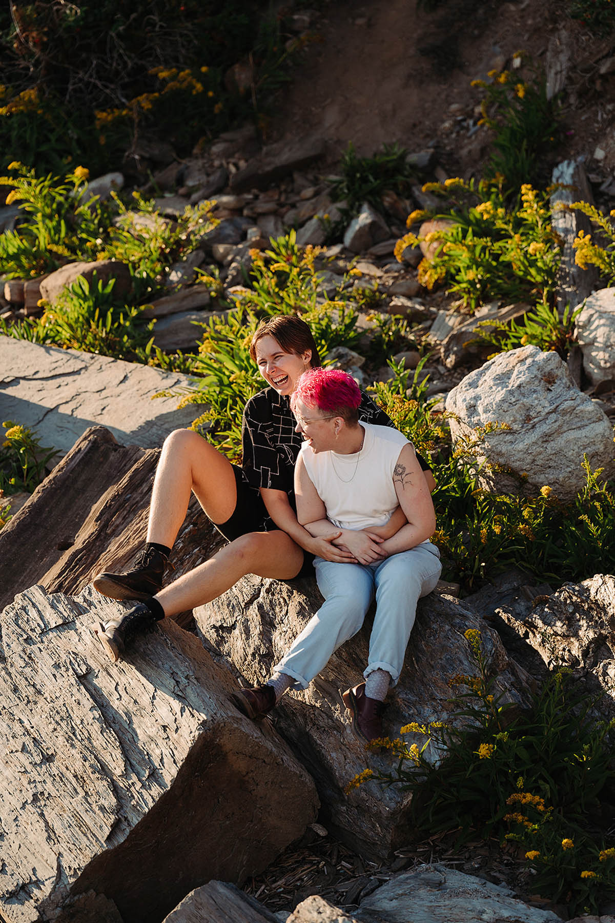 Two white people sit on a large rock covered in green patches of grass. The people are embracing and laughing. The one in the front has pink hair, and the one in the back is brunette. 