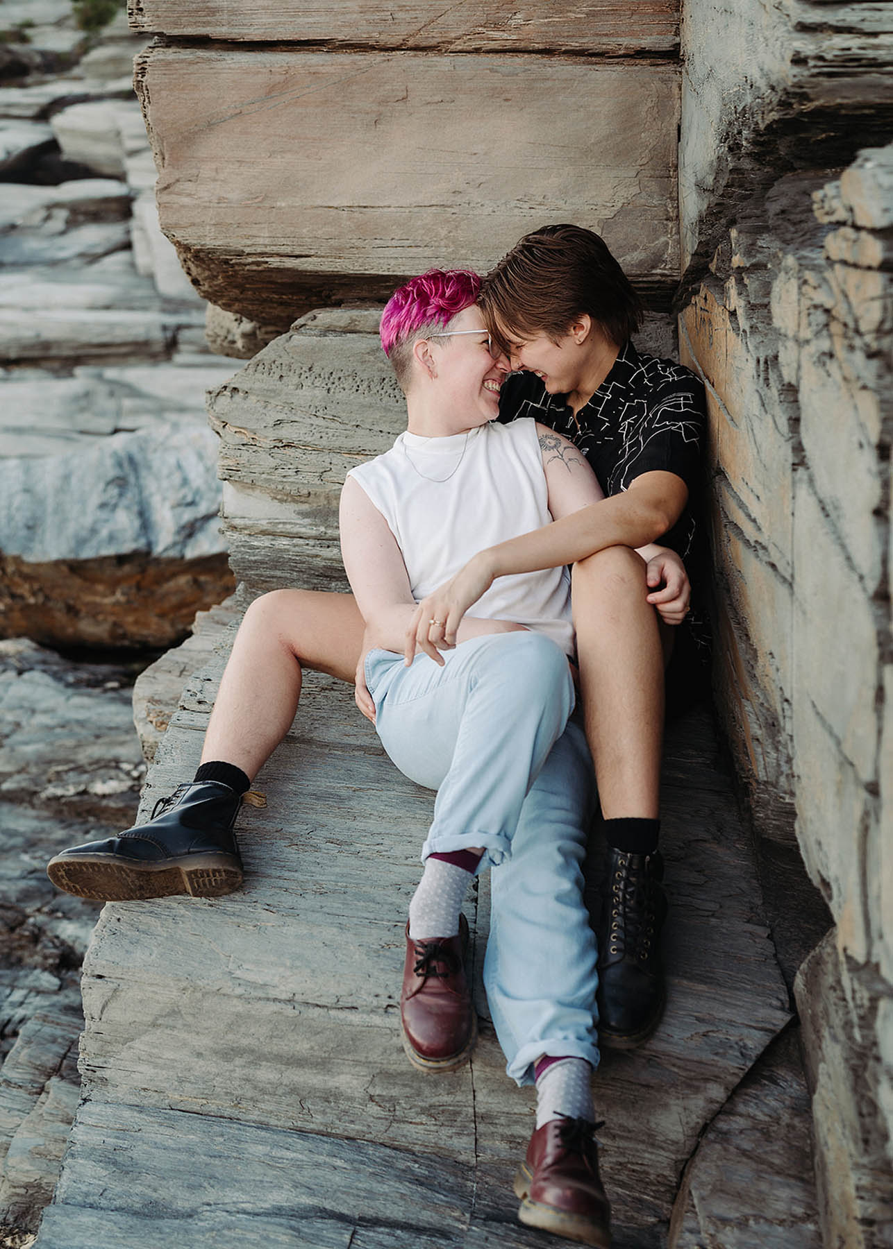 Two white people sit on the ledge of a large rock, with their foreheads touching. They are both wearing Doc Marten boots. 