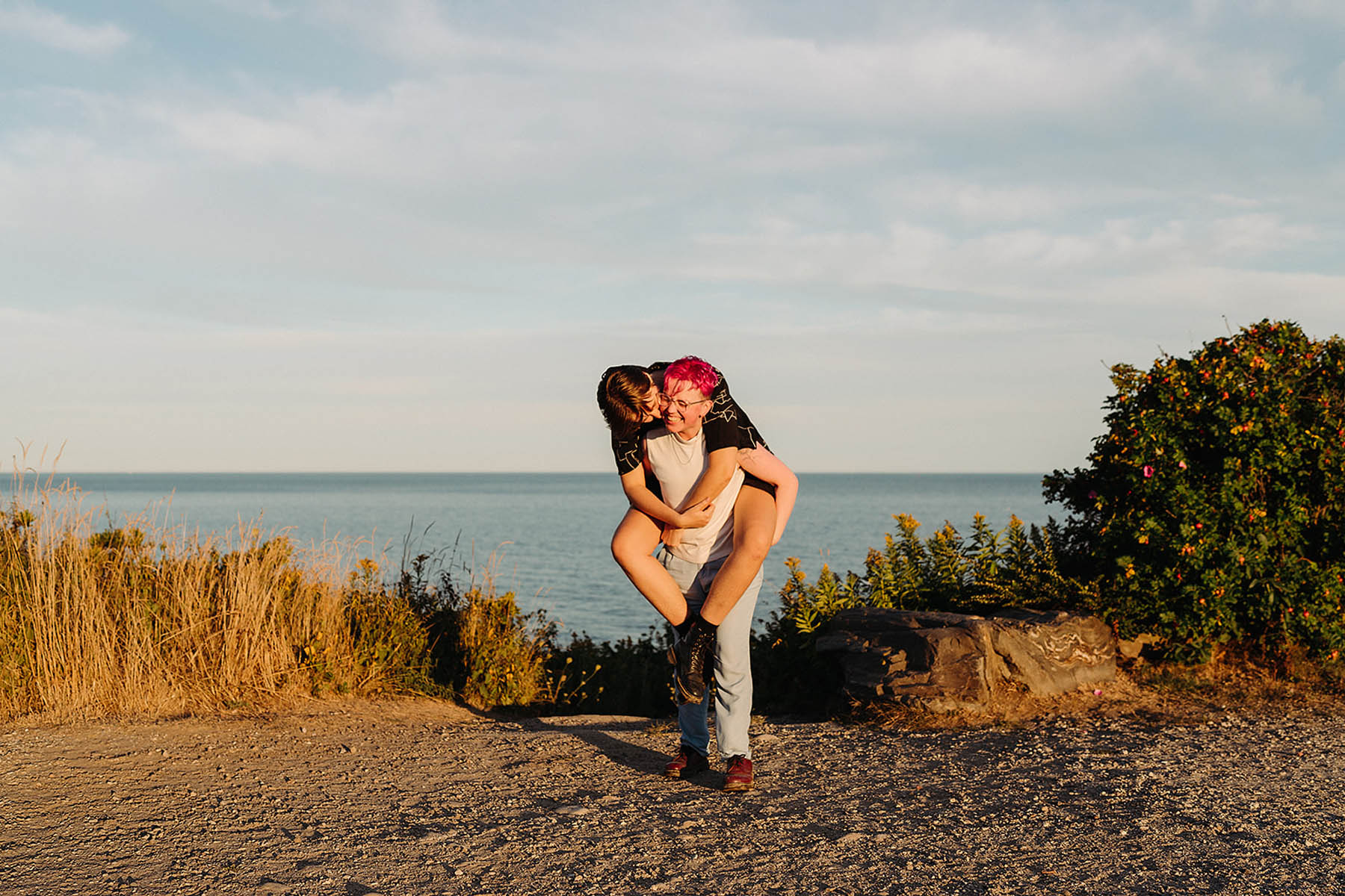 Two white people are in front of the ocean. One person has pink hair and a white tank top. The other person has brown hair and is wearing a black button-up shirt. They are carrying the other person on their back. 