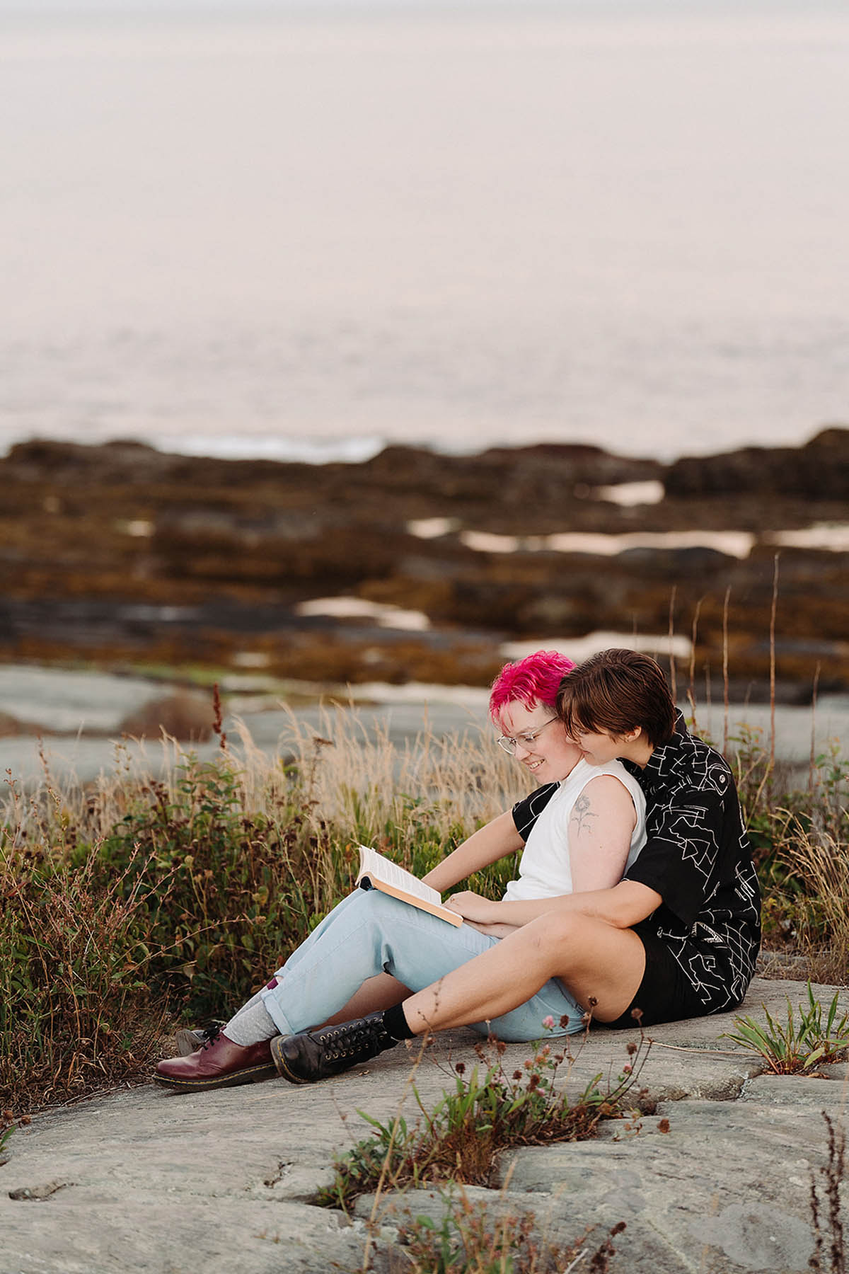 Two white people sit on a large rock with wild grass in the background behind them. One has pink hair and is holding a book open in front of them. The other person sits behind them and reads over their shoulder. 