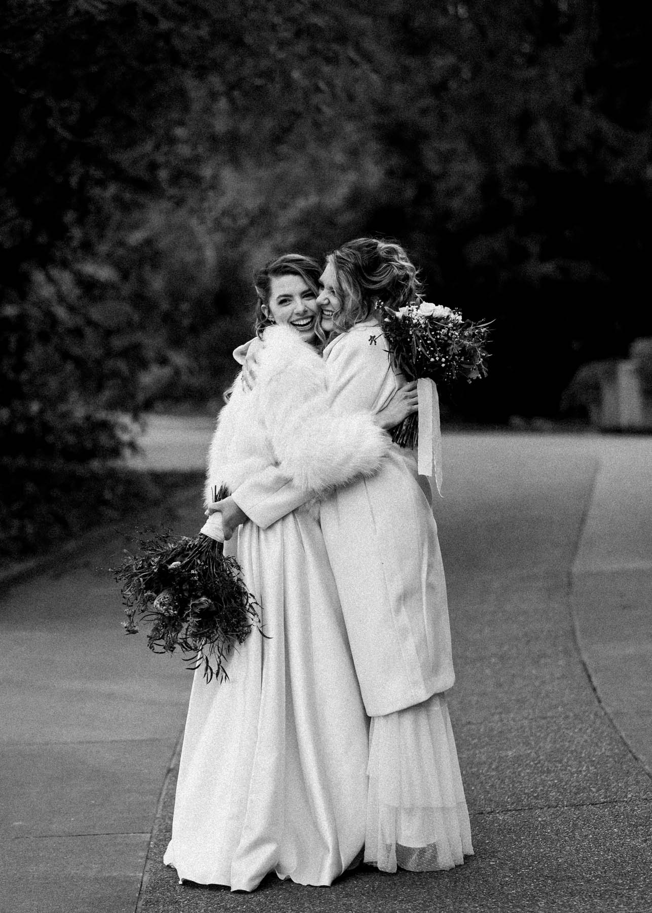 A black-and-white photo of two brides hugging. They are both wearing long white jackets.