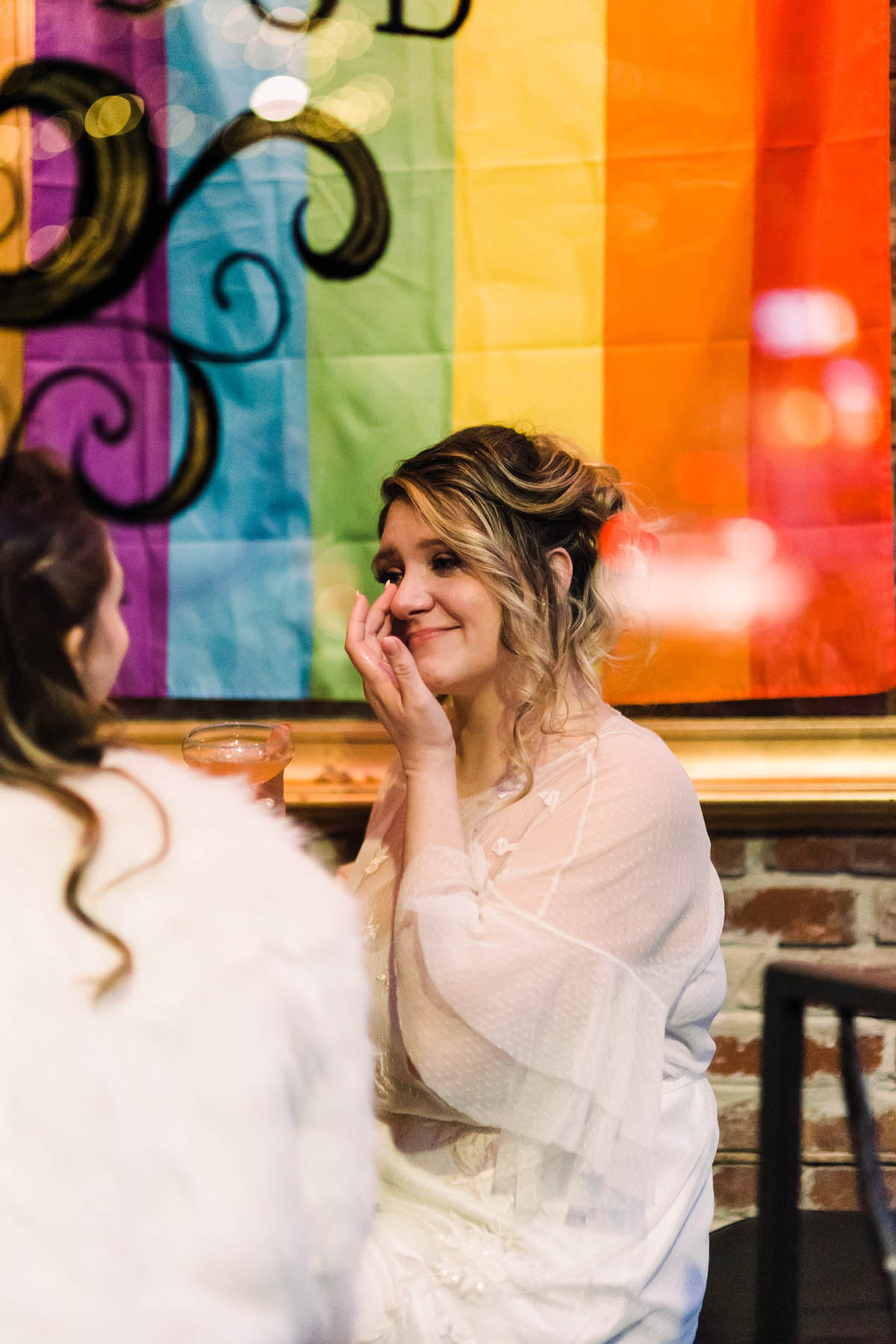 The couple sits at a bar in front of a rainbow Pride flag. One of them is wiping a tear from her eye.