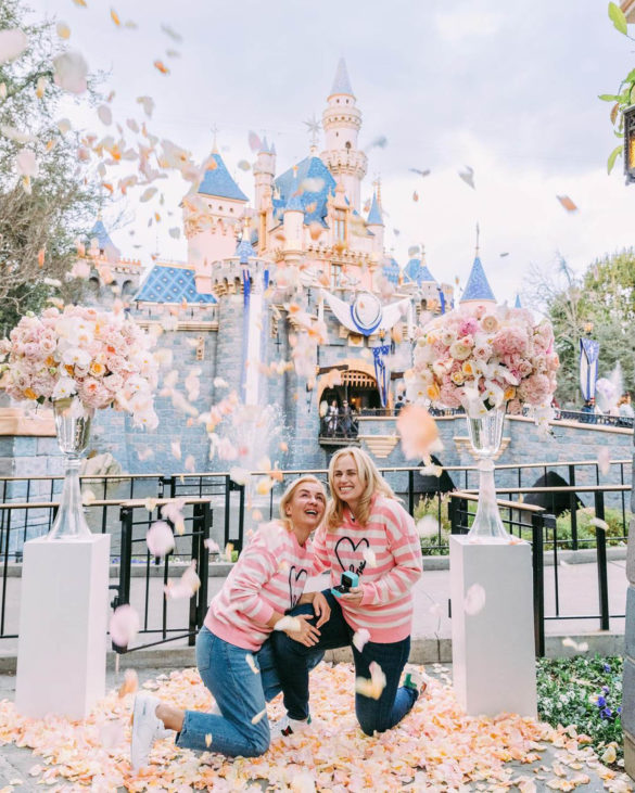 Rebel Wilson and her partner Ramona kneel in front of Sleeping Beauty's castle in Disneyland. They are wearing matching sweaters and smiling.