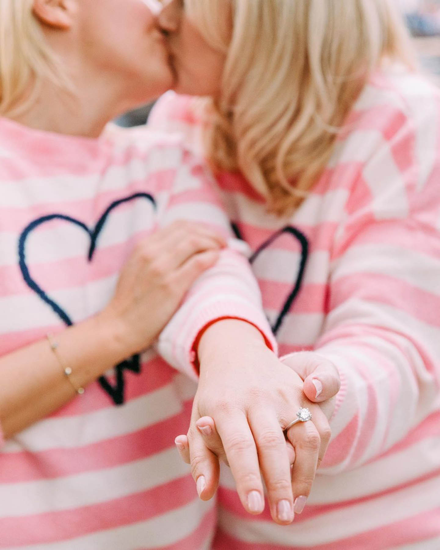 Two white women in matching sweaters kiss and show off a Tiffany engagement ring. 