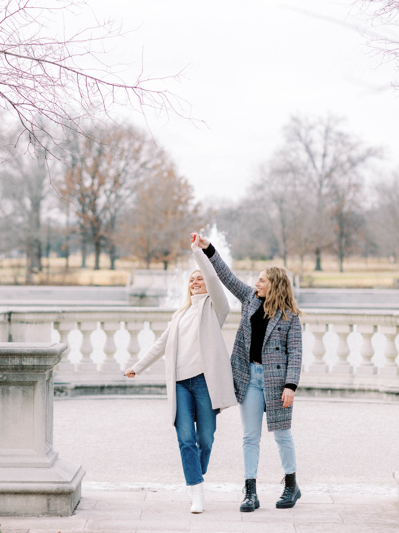 Two white people with blonde hair hold hands as one of them twirls in front of a marble and concrete fountain.