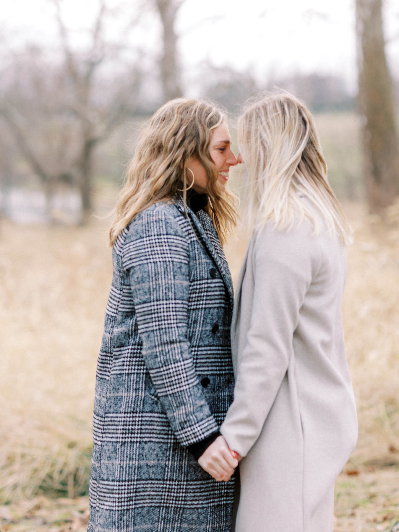 Two blonde people hold hands and touch noses with a winter wooded background behind them.