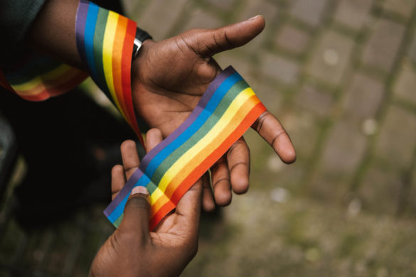 A rainbow ribbon is being held by a black person, whose hands are the only thing we can see besides the rainbow ribbon.