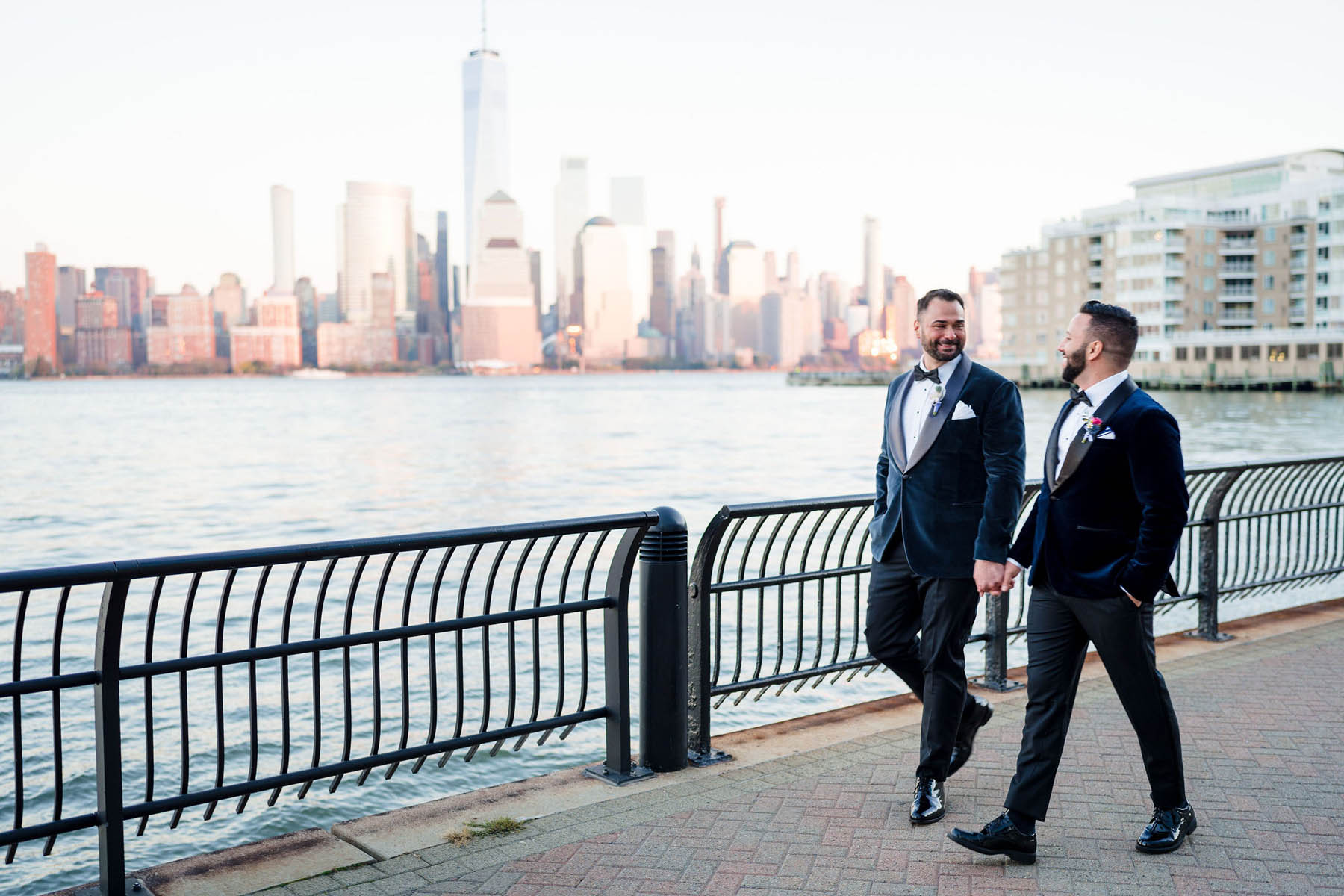 Two grooms walk hand-in-hand along the Hudson River on a sunny afternoon.