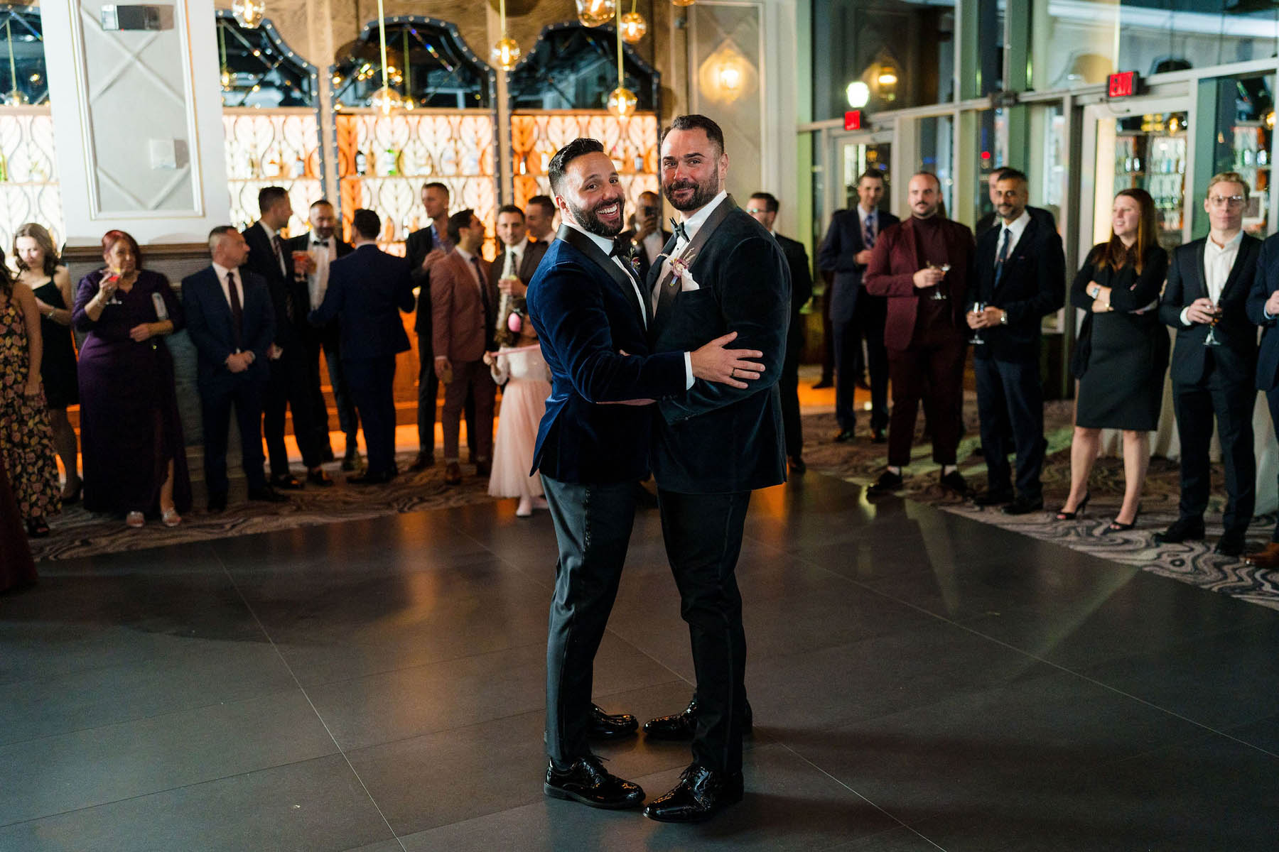 The grooms stand in the middle of the dance floor and smile at the camera.