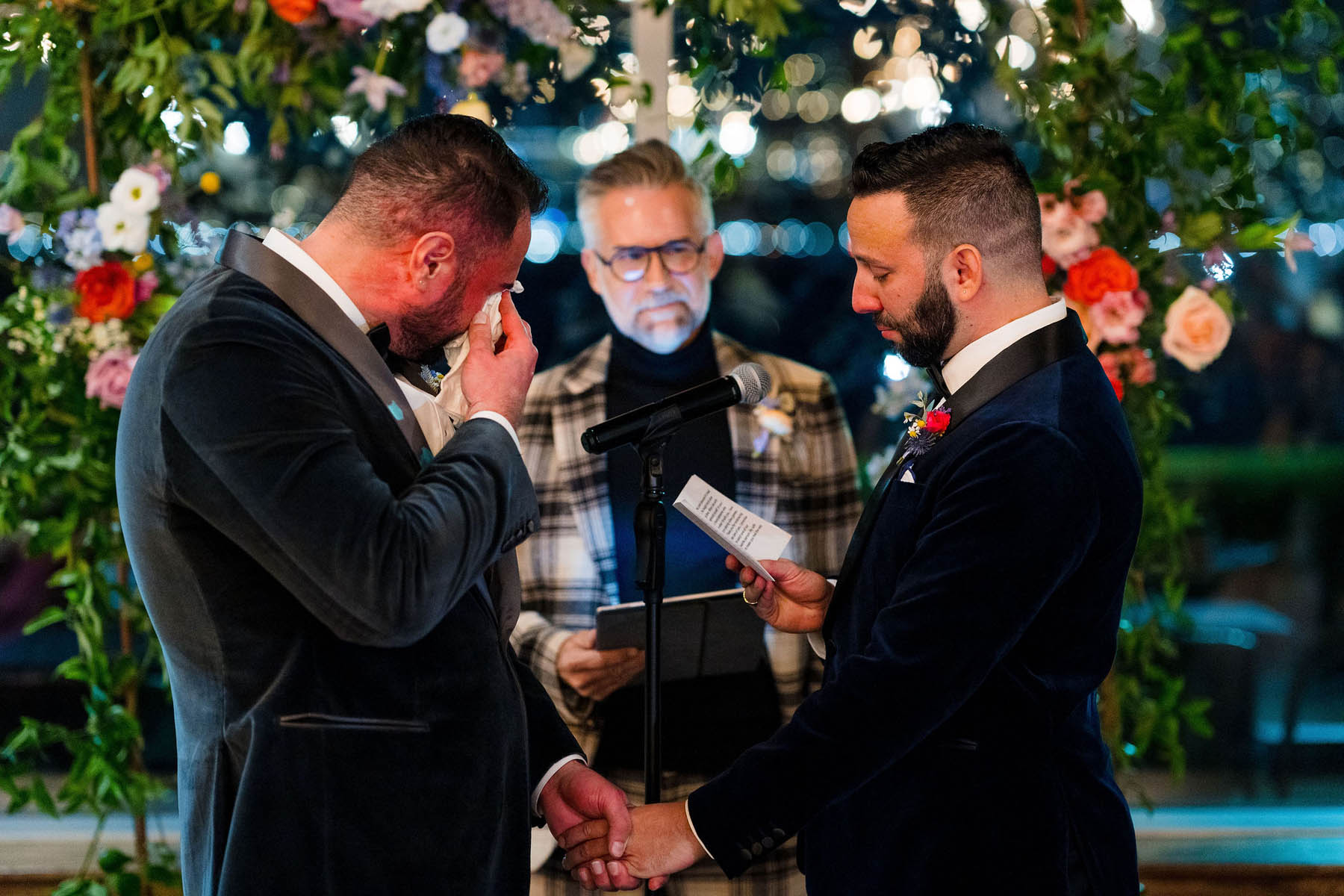 The grooms stand at the altar and one reads his vows. The other dabs his eye with a tissue.