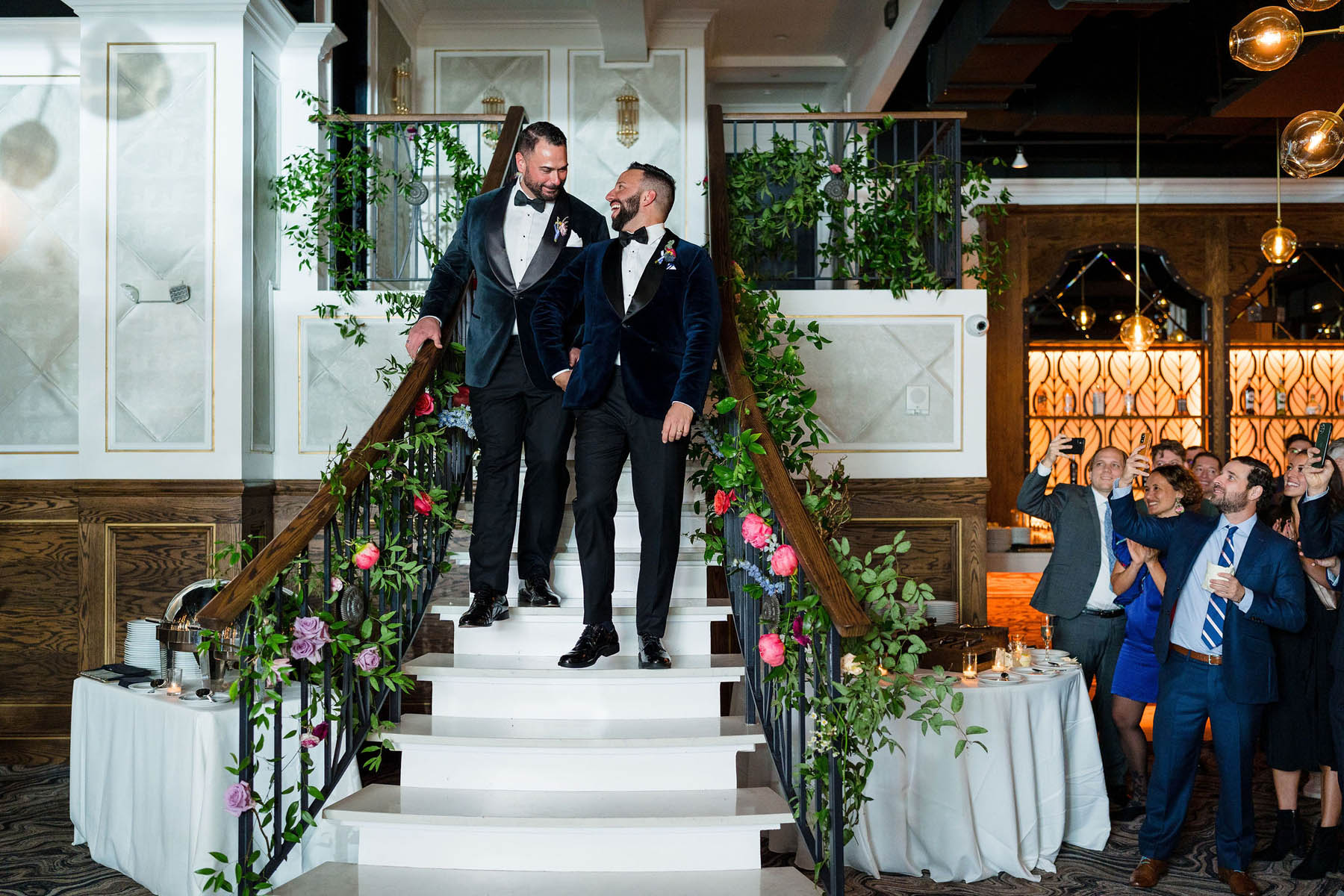 Two grooms walk down a set of stairs. They are smiling at each other.