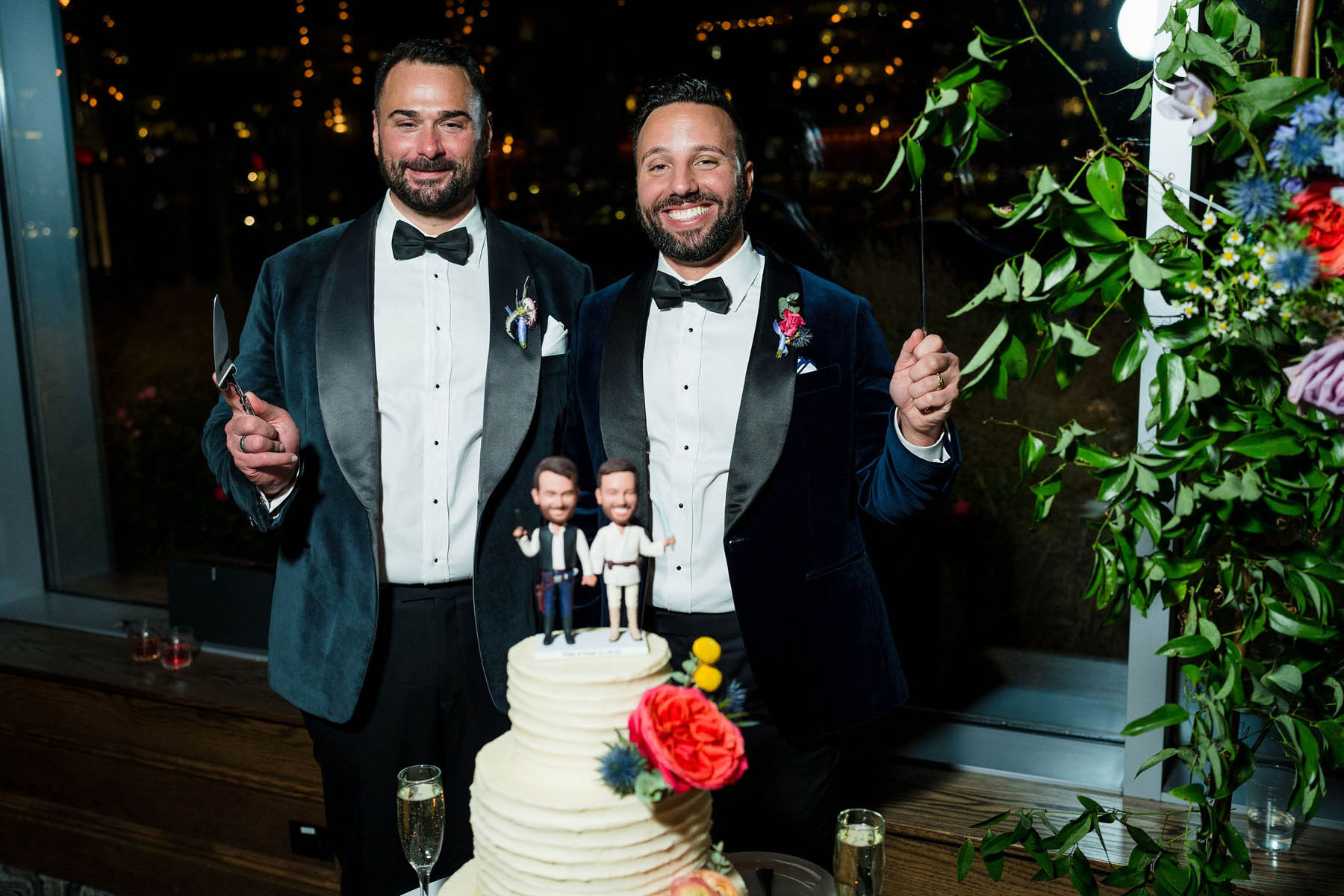The grooms stand behind their wedding cake, which has a Star Wars cake topper on it that looks like them. They pose like the figurines, smiling.