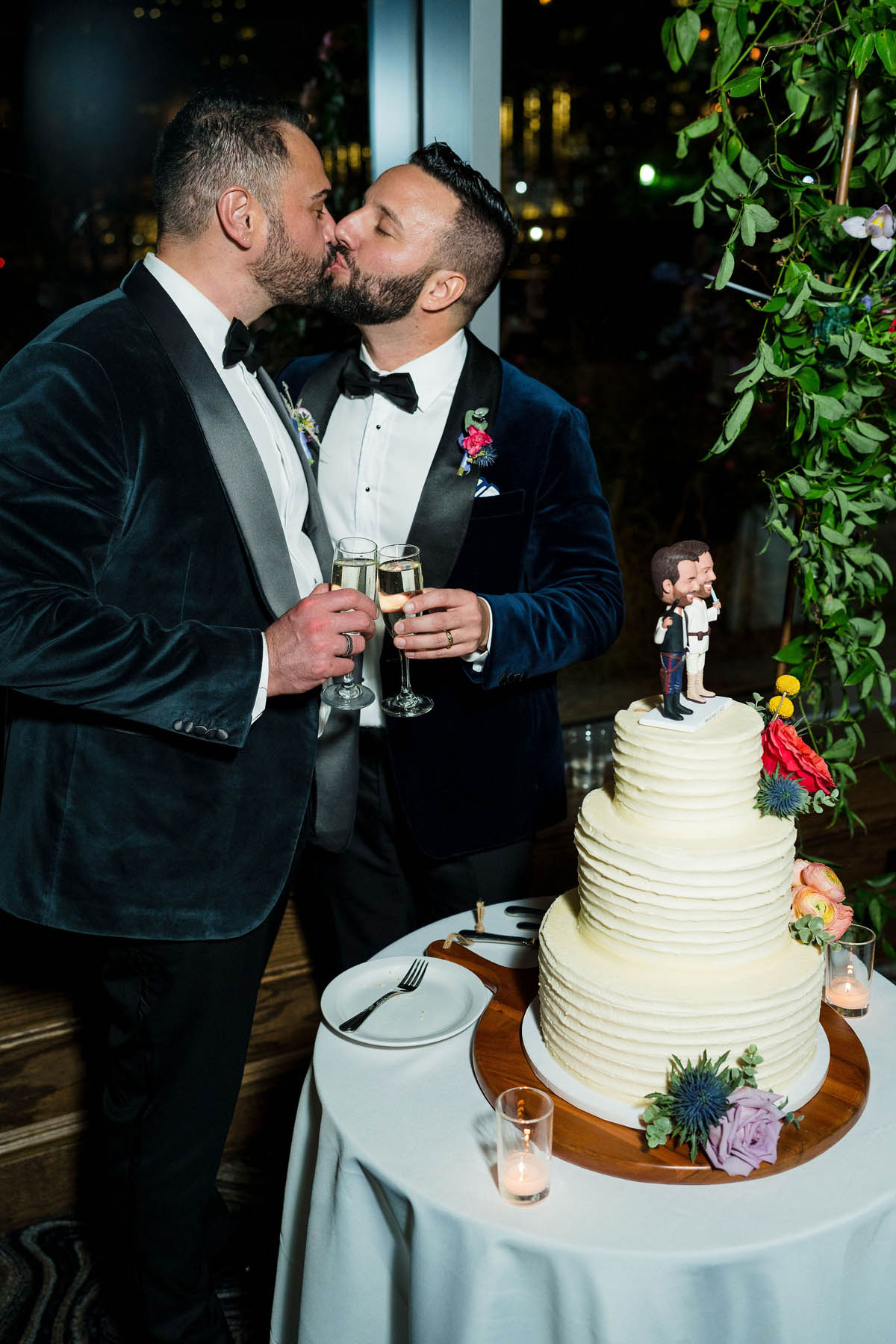 The grooms share a kiss in front of their wedding cake as they clink their champagne glasses.
