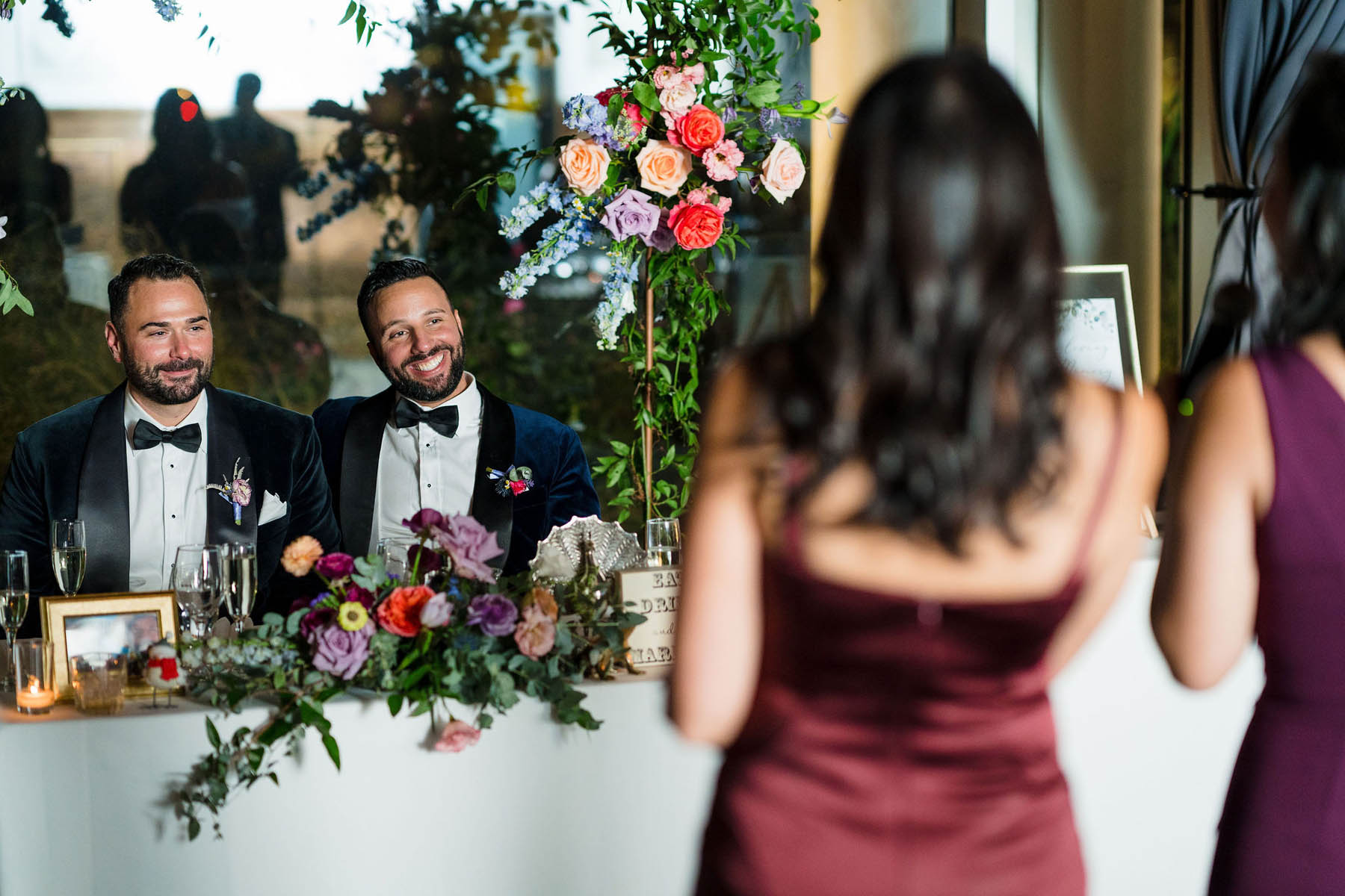 The grooms sit at a sweetheart table with floral decor as two people in burgundy dresses give a speech.