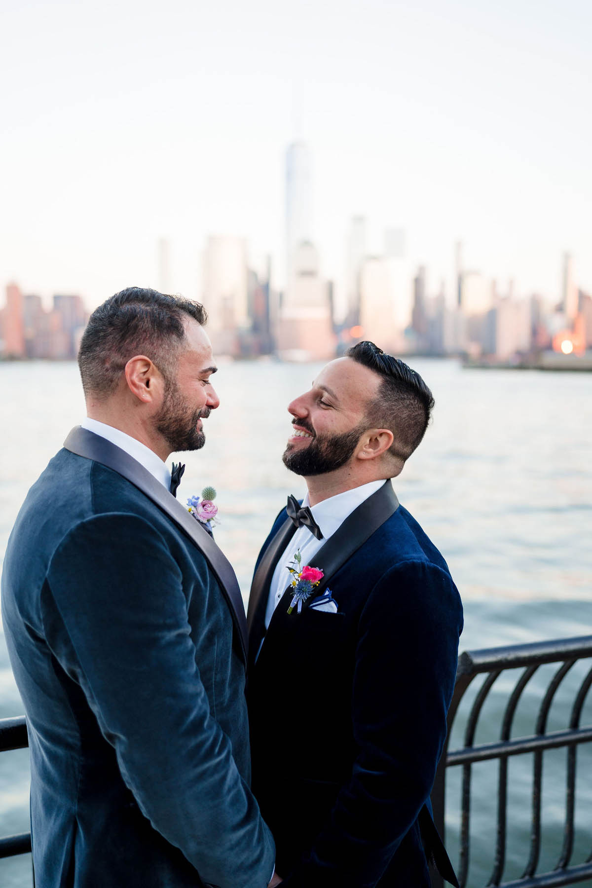 Two grooms stand in front of the downtown Manhattan skyline. They are wearing tuxes and smiling at each other