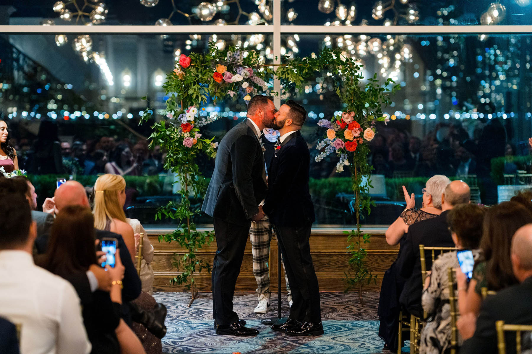Two grooms stand in front of a set of large windows and a floral arbor. They share a kiss.