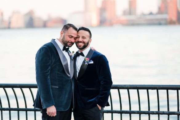 Two white grooms stand in front of the Hudson River, arms around each others' backs and smiling.