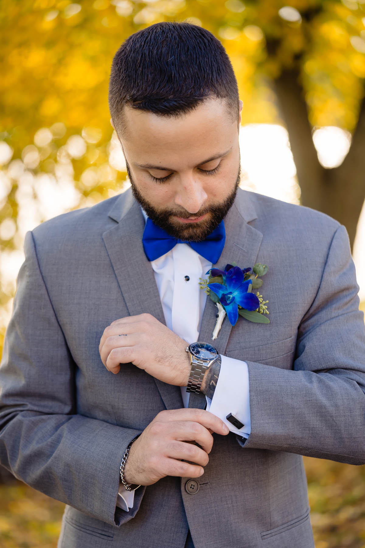 A person with short brown hair and a beard adjusts his cuff links. He is wearing a gray suit with blue tie and boutenniere. 
