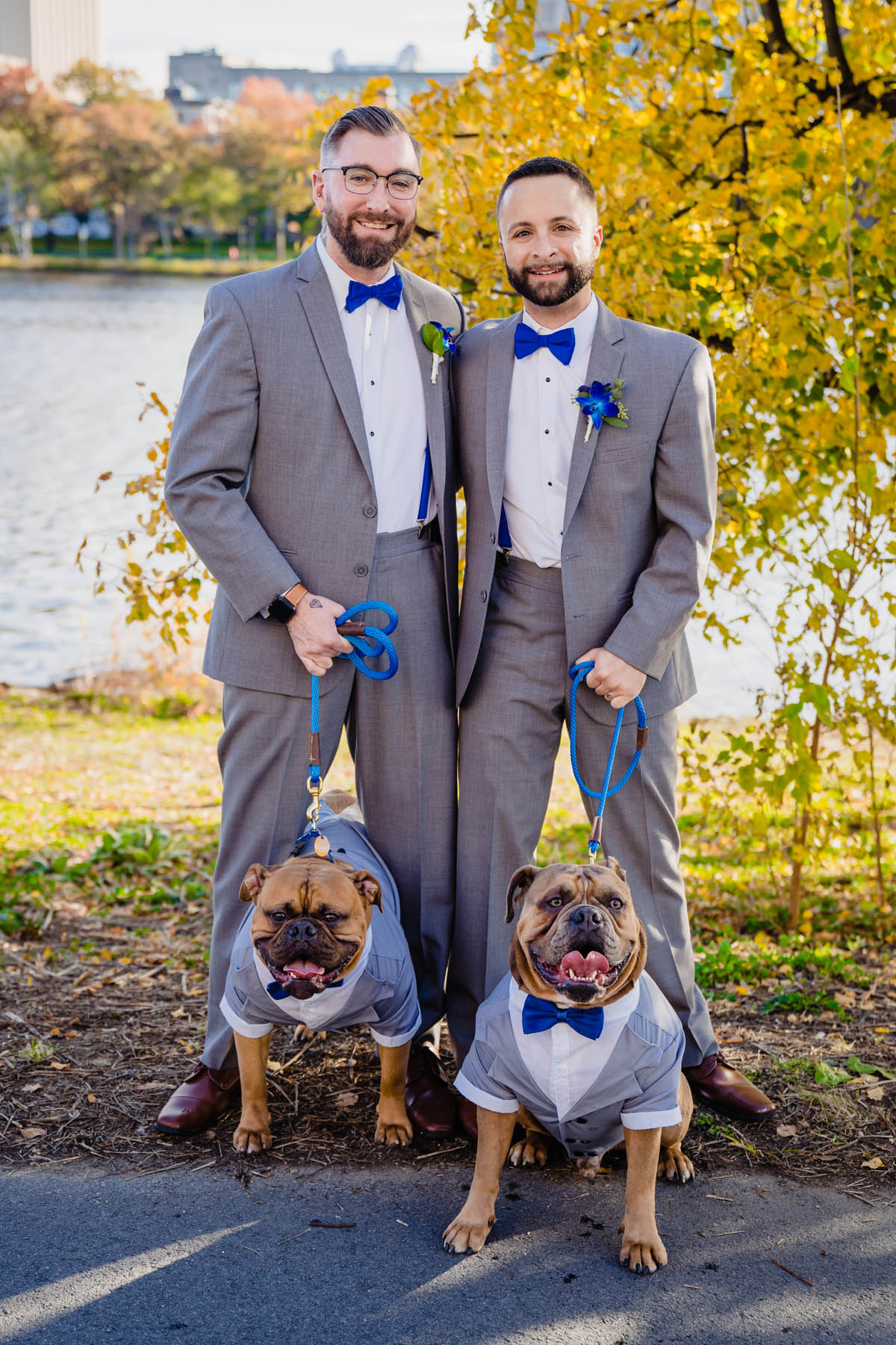 Two dogs and their owners all stand in matching gray suits, smiling at the camera