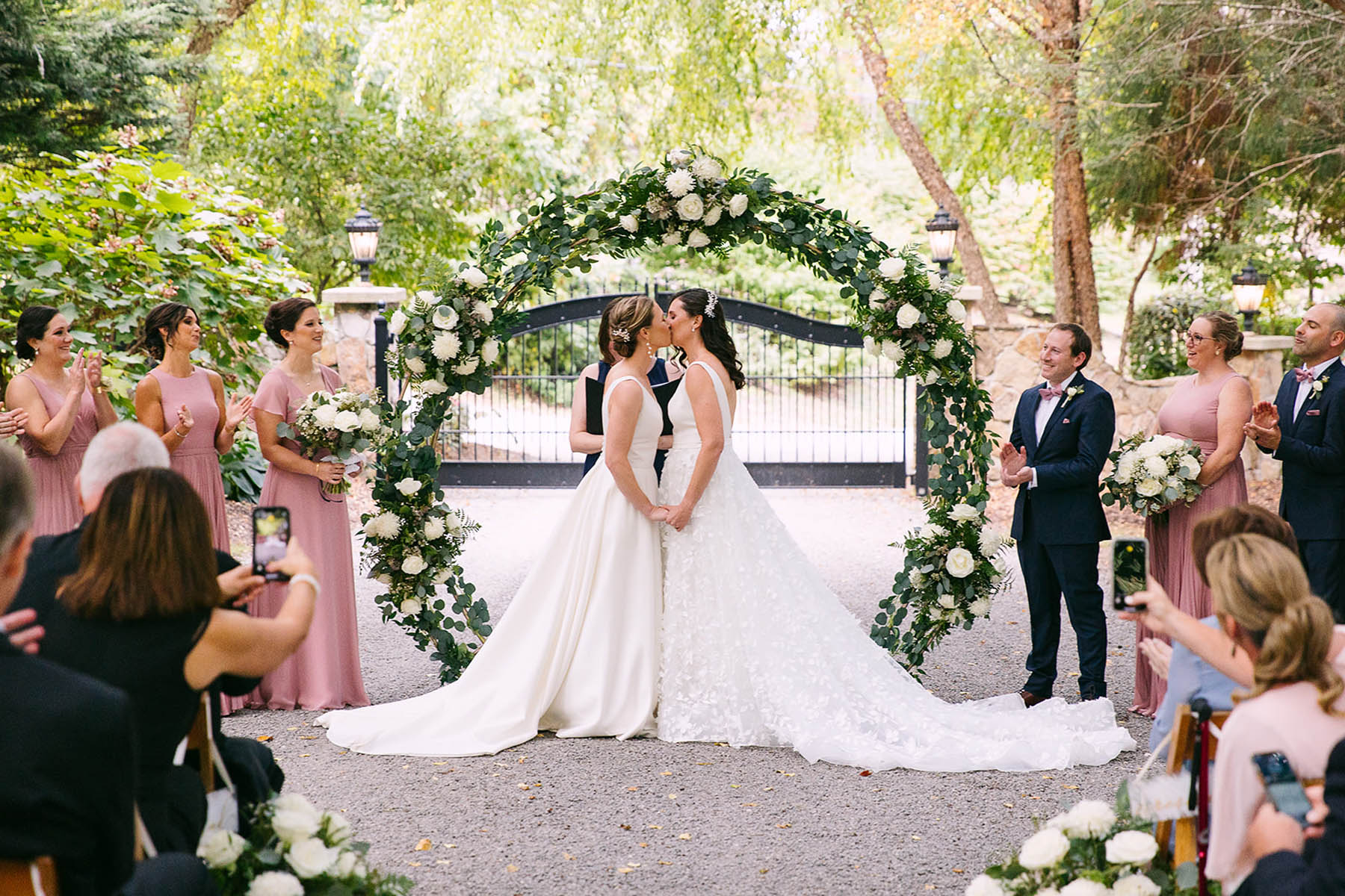 Two brides, one brunette and one blonde, stand in front of a round floral arbor, kissing.