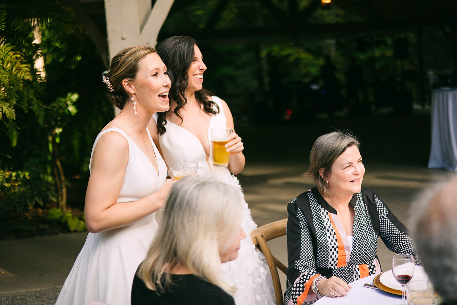 Two brides stand and laugh as someone gives a toast. 