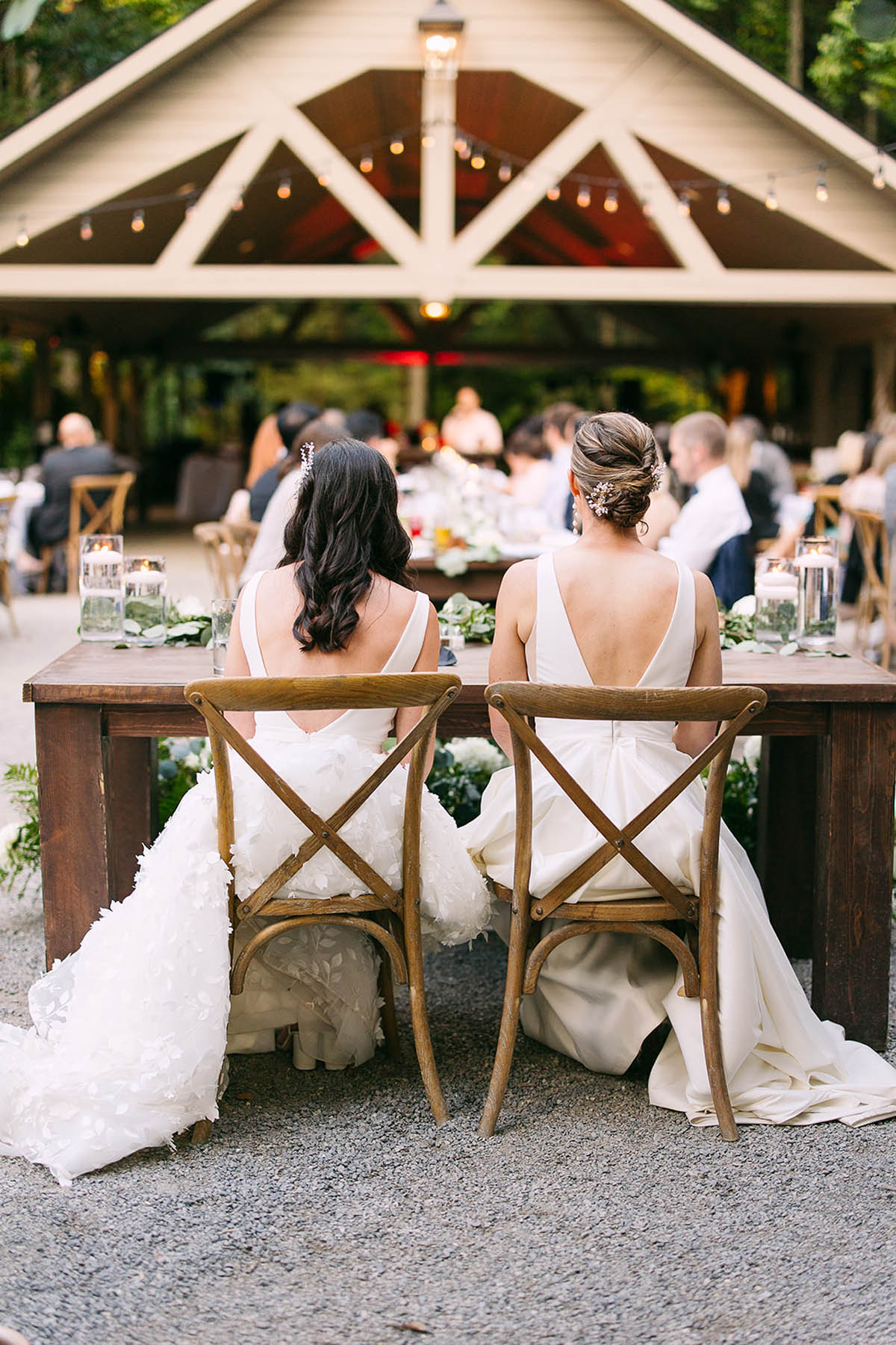 Two brides sit at a sweetheart table in front of a barn-like structure at their wedding reception.