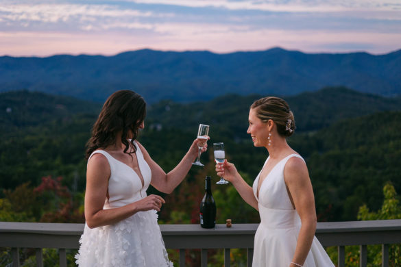 Two white brides stand on a wooden balcony in front of a North Carolina landscape. They have prosecco glasses and are smiling at each other.
