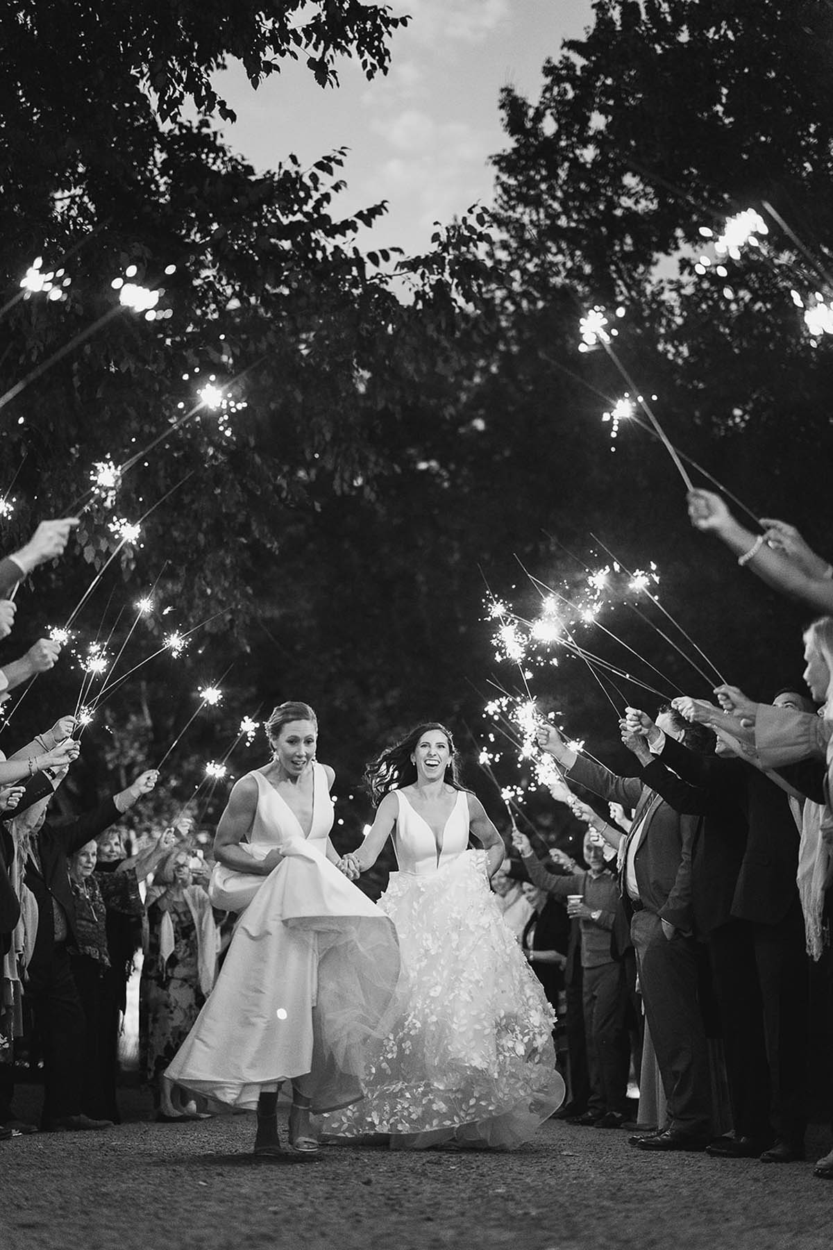 A black-and-white photo of two brides running down an aisle of friends and family holding sparklers.