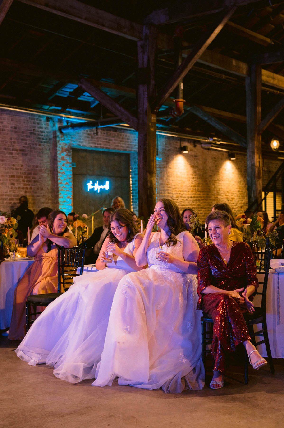 The newlyweds sit and laugh as someone gives a toast. They are in a brick and wooden loft building.