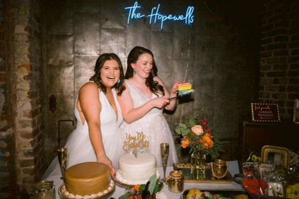 Two white brides stand behind a dessert table with a slice of rainbow cake.