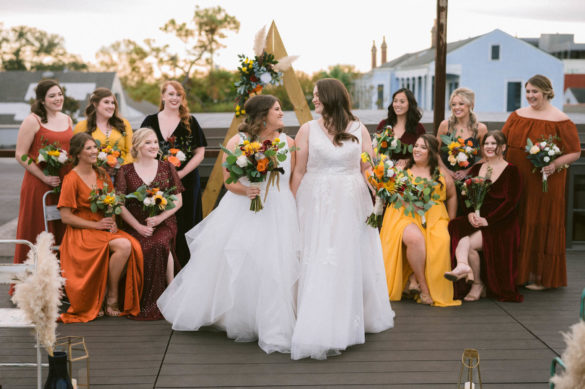 Two white people with long brown hair and white gowns stand in front of an angled arbor. They are surrounded by friends wearing vibrant colors like marigold and burnt sienna.