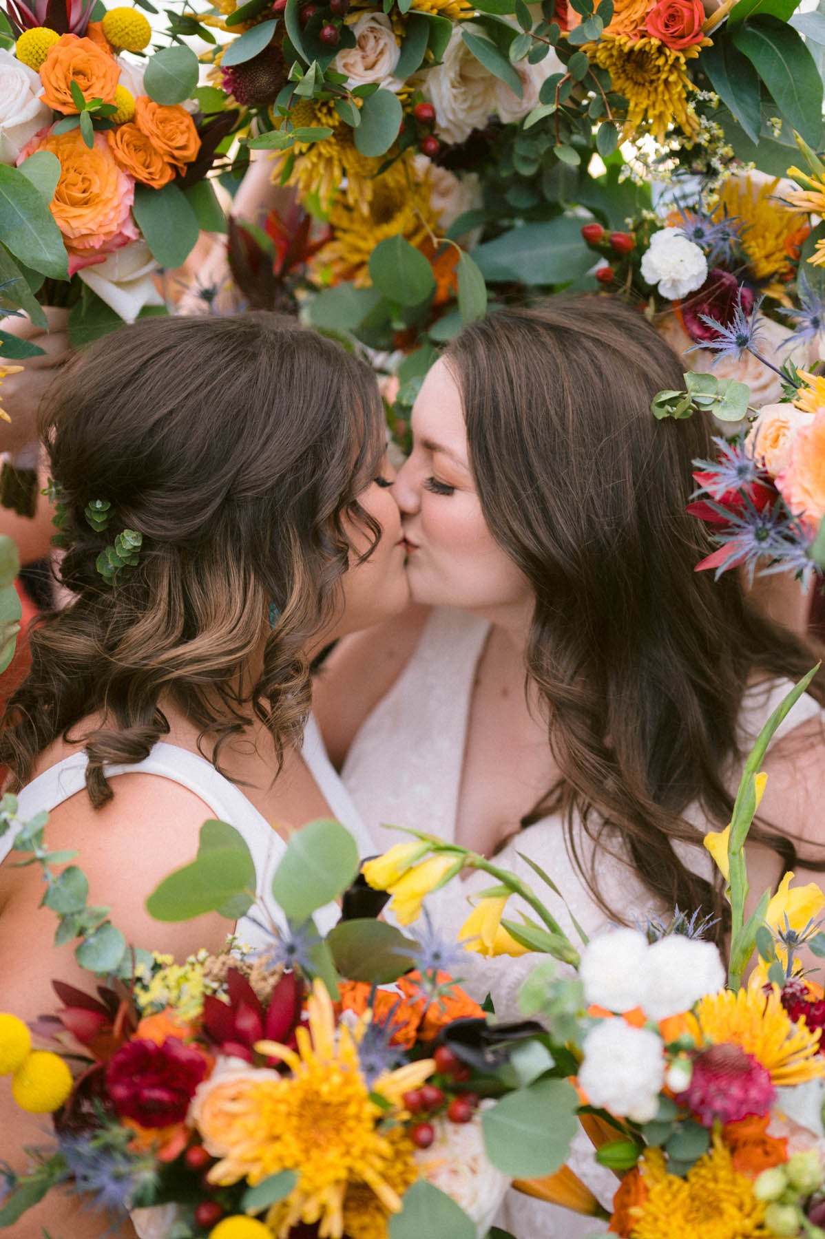 Two white people with brown hair kiss, surrounded by bouquets of vivid jewel tone flowers.