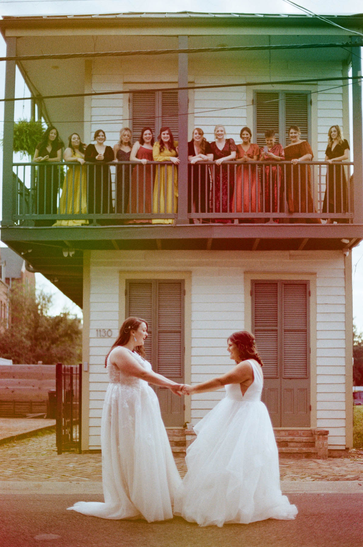 Two white people in long white gowns hold hands and look at each other. On a balcony above them, their wedding party is smiling down at them. 