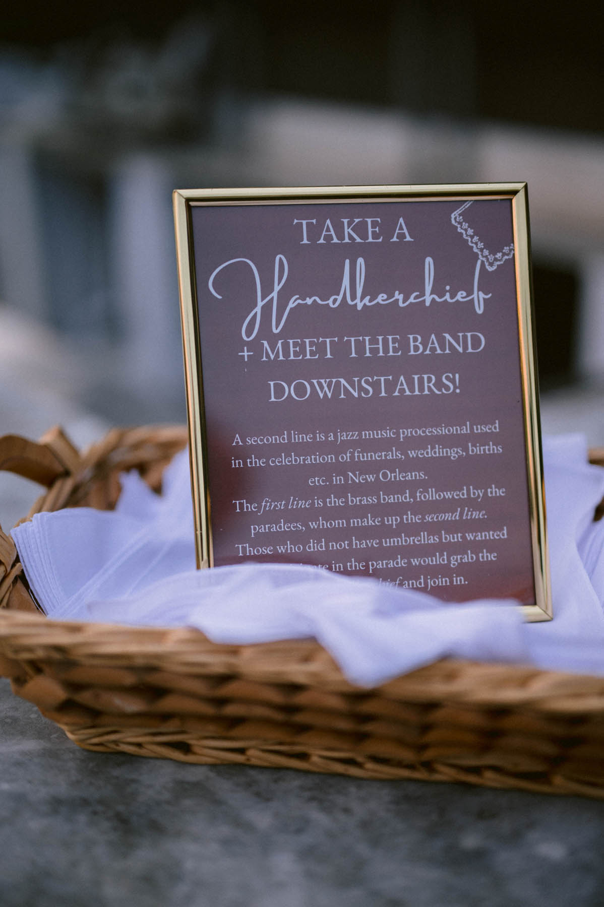 A gold frame sits in a basket of white handkerchief. The text inside reads: Take a handkerchief + meet the band downstairs! A second line is a jazz music processional used in the celebration of funerals, weddings, births, etc. in New Orleans. The first line is the brand band, followed by the paradees, whom make up the second line. Those who did not have umbrellas but wanted to participate in the parade would grab their handkerchief and join in. 
