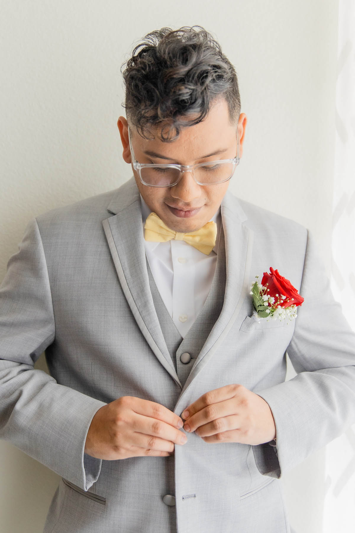 A Latinx groom buttons the top button of his gray suit. He is wearing clear frame glasses and a yellow bowtie.