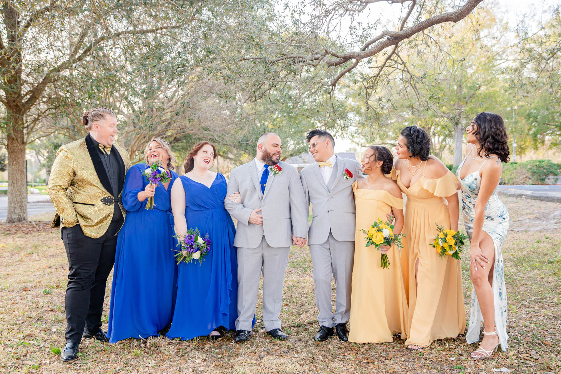Two Latinx grooms in blue suits stand in the center of a line of their wedding party. Everyone is laughing, and they are standing in front of a backdrop of trees.