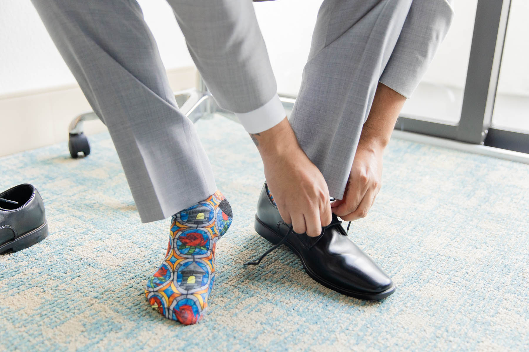 A groom puts on a shoe. On his other foot, he is wearing a sock that is designed like stained glass.