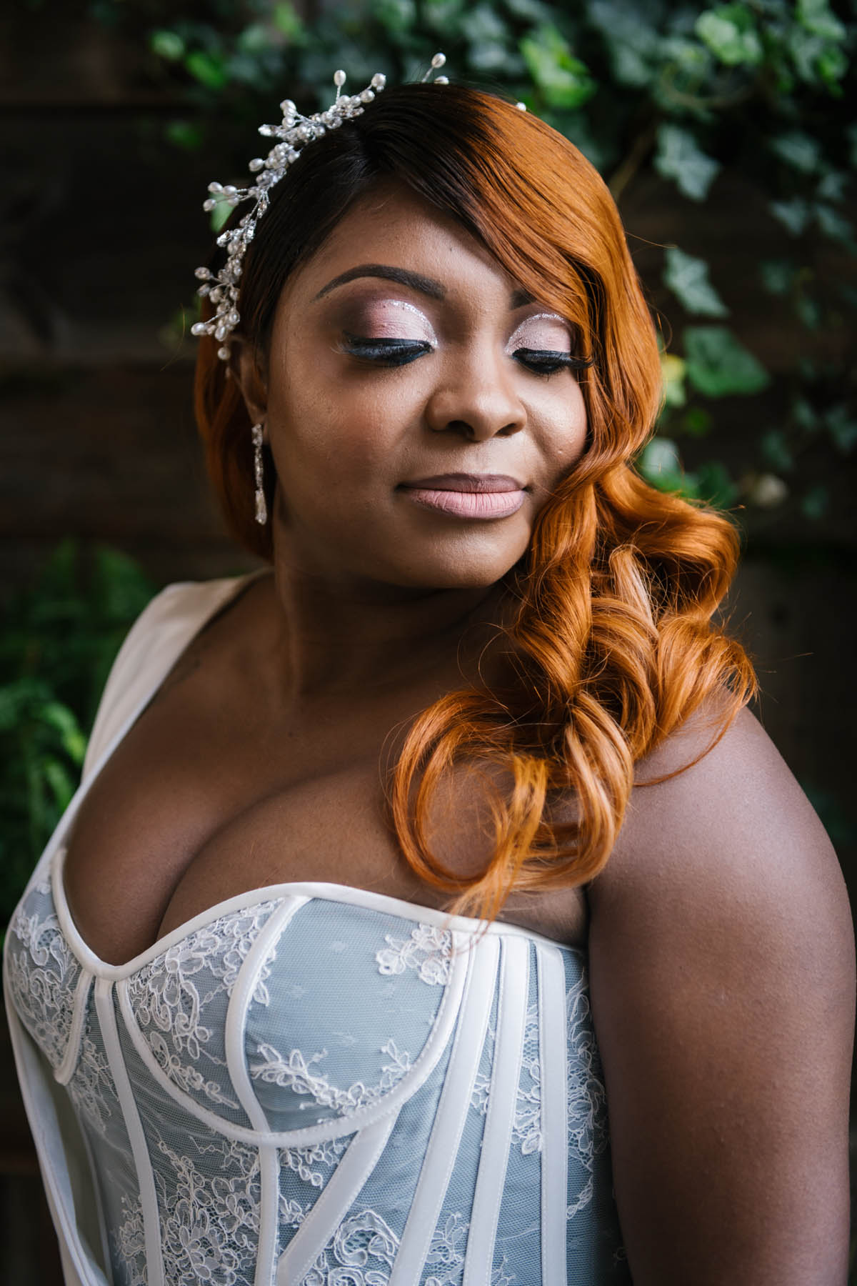 A Black bride with a fitted corset and ombre auburn hair looks down over her shoulder, showing off loose Hollywood waves and a rhinestone and pearl pinned hairpiece on her right side.