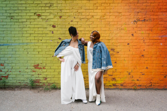 Two Black brides stand in front of a rainbow mural wall wearing white attire, jean jackets and Pride sneakers