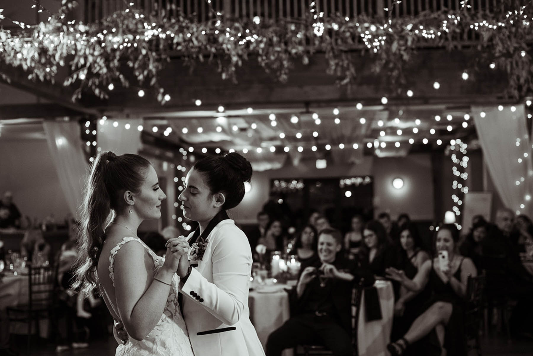A black-and-white photo of two brides dancing. One has a sleek bun and the other has a slicked back ponytail with cascading curls.