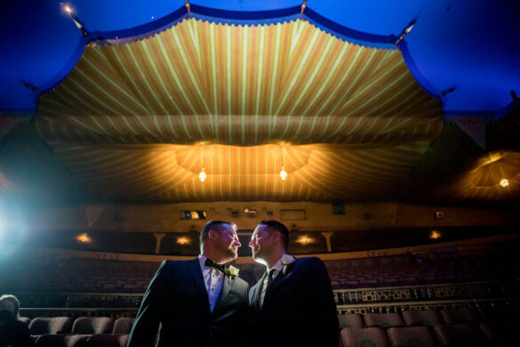 Two white grooms in suits stand on a stage with blue curtains, with the house lights of a theatre behind them.