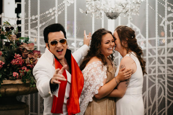 Two white marriers in white and gold attire stand behind an Elvis impersonator as they smile and putheir heads together.