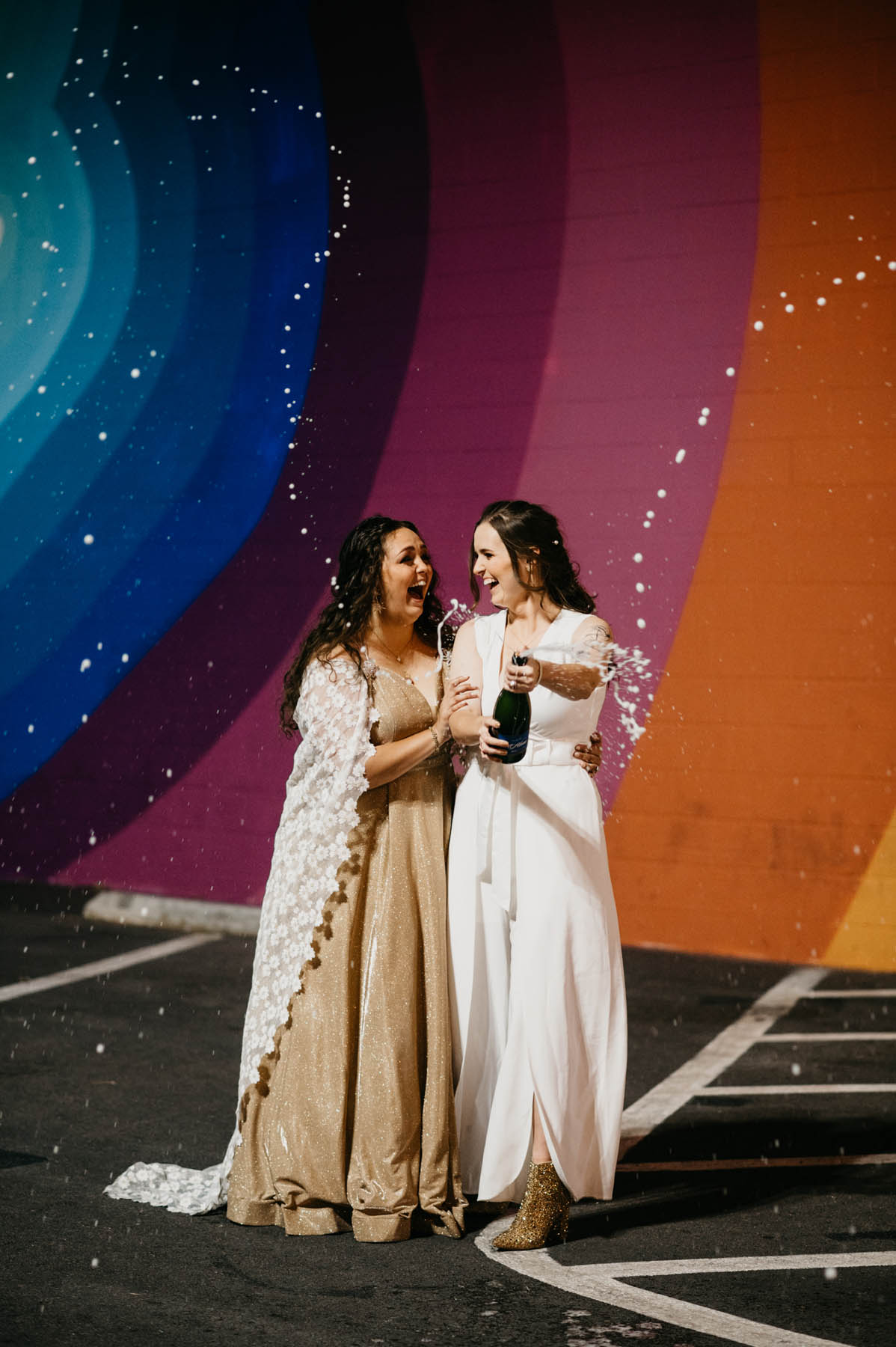 The newlyweds, both brunettes, stand in front of a rainbow color mural and pop a bottle of champagne.