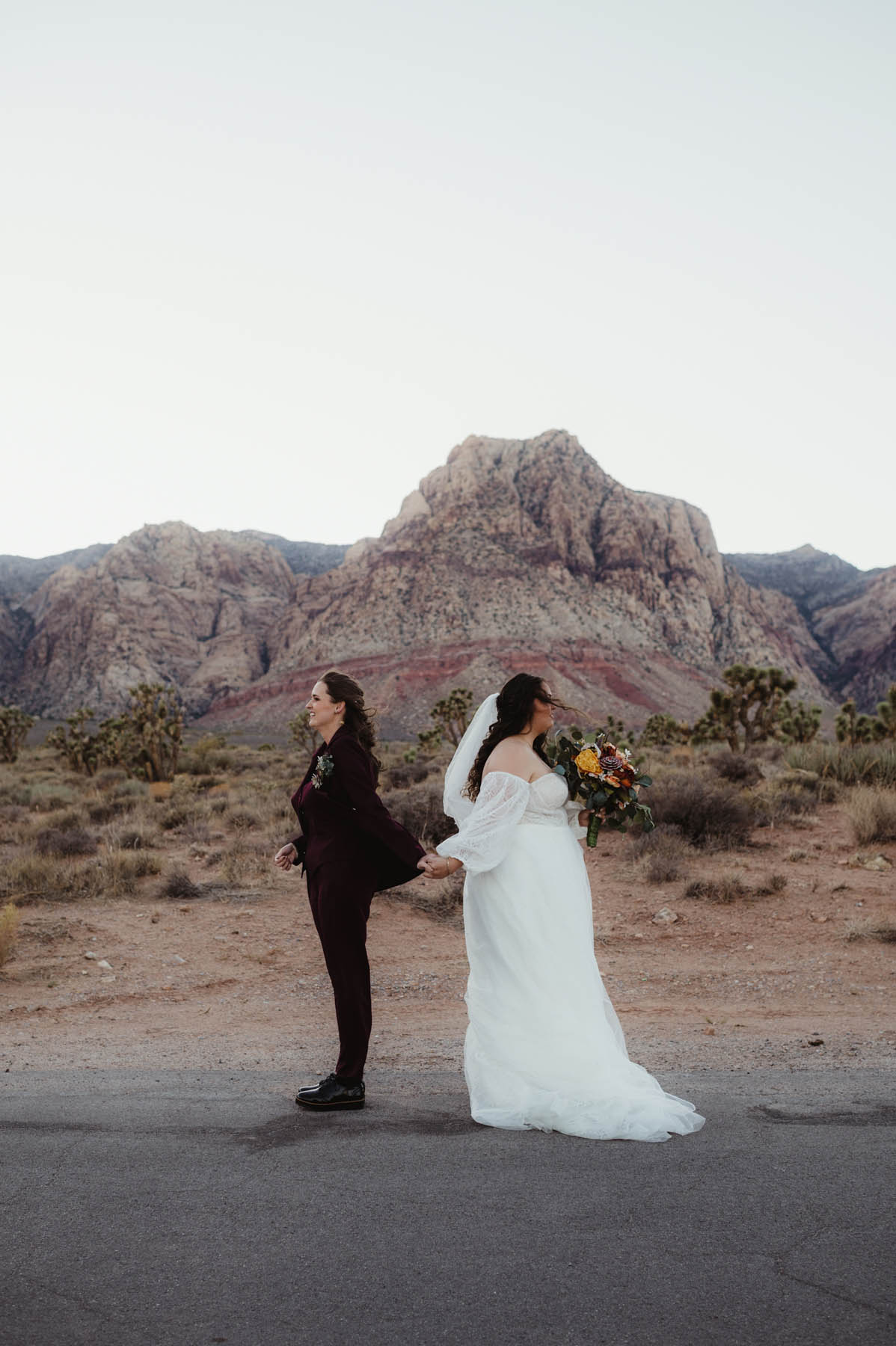 Two whie brunettes hold hands and stand back to back against a rocky desert background.