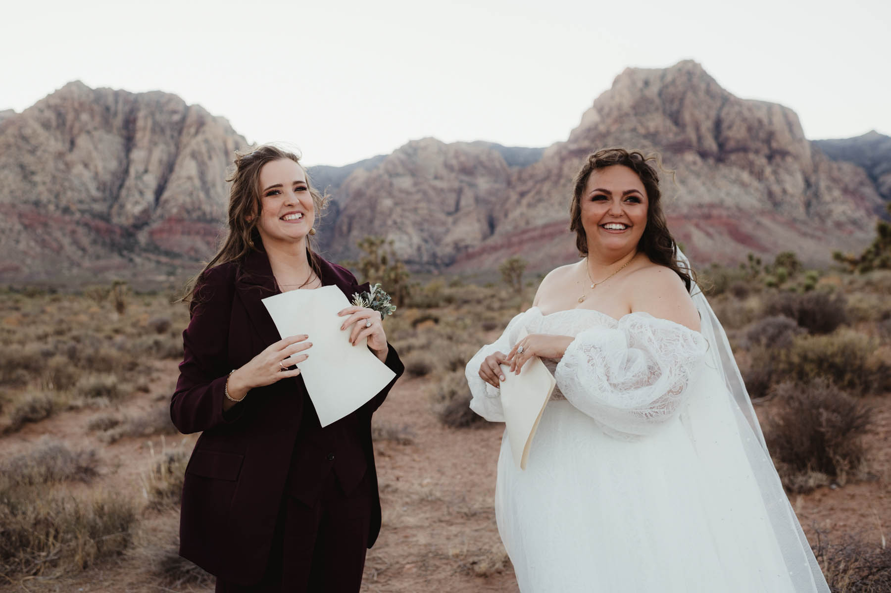 Two white brunettes stand in a desert with their vows written on paper in their hands.